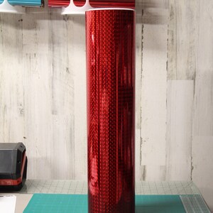 May include: A roll of red holographic vinyl with a textured, speckled surface. The vinyl is rolled up and sitting on a green cutting mat.