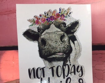 May include: A white sticker with a black and white cow wearing a flower crown. The cow has a glittery finish. The text "Not Today Heifer" is written in black below the cow.
