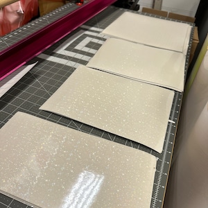 May include: Four rectangular sheets of light gray material with a sparkling, glittery finish are arranged on a gray cutting mat. A pink metal bar and a wooden dowel are visible in the background, suggesting a crafting or production setting.