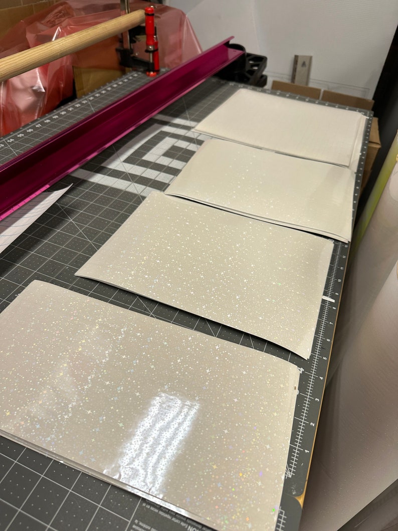May include: Four rectangular sheets of light gray material with a sparkling, glittery finish are arranged on a gray cutting mat. A pink metal bar and a wooden dowel are visible in the background, suggesting a crafting or production setting.