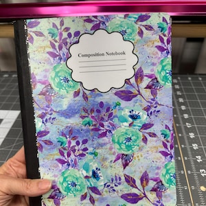 May include: A composition notebook with a floral design. The cover features teal and purple flowers with gold accents on a light blue background. The notebook has a black spine and the text "Composition Notebook" on a white label.