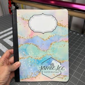 May include: A notebook with a colorful watercolor design featuring blue, pink, and gold glitter accents. The cover includes a white label area and the text "jamie lee CUSTOM BOUTIQUE" in a black hexagon.
