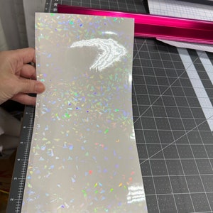 May include: A sheet of iridescent white vinyl with a holographic finish. The vinyl has a scattered pattern of small, iridescent triangles.