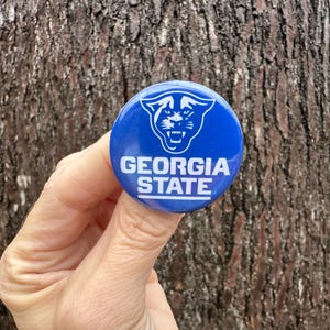 May include: A round, blue button featuring the Georgia State University logo. The logo is a white panther head with the words "GEORGIA STATE" in white block letters. The button is held in front of a tree trunk.