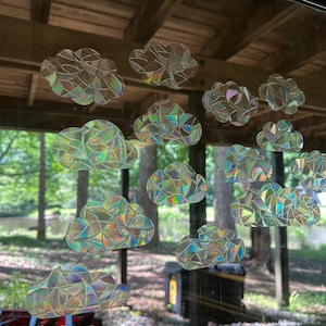 May include: A collection of iridescent cloud-shaped window clings. The clings are made of a mosaic of small, reflective pieces that create a rainbow effect. The clouds are arranged on a window with a natural outdoor background.