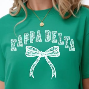 May include: A bright green short-sleeved t-shirt featuring the text Kappa Delta in a white floral lace pattern. Below the text is a large white bow graphic also filled with a detailed floral lace design. The shirt has a crew neckline.