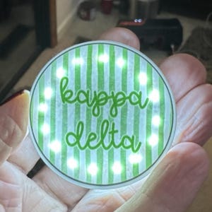 May include: A round, illuminated Kappa Delta emblem. The design features a green and white striped background with the words "kappa delta" in green script. The emblem is surrounded by small, bright lights.