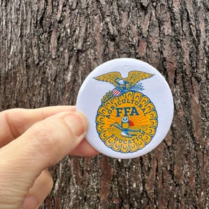 May include: A white button with the FFA emblem, featuring a golden eagle, a blue and gold design, and the text "AGRICULTURAL EDUCATION FFA". The button is round and has a small hole at the top.
