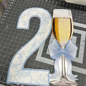 May include: A large, decorative number "2" and a champagne glass cutout. The number is white with a blue floral pattern and a blue border. The champagne glass is filled with bubbly liquid and has a blue bow.