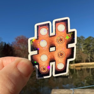 May include: A sticker of a brown hashtag symbol with white circles inside. The sticker has a holographic finish with iridescent sparkles.