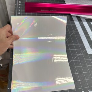 May include: A sheet of iridescent white vinyl with a holographic finish. The vinyl is lying flat on a black cutting mat with a ruler.
