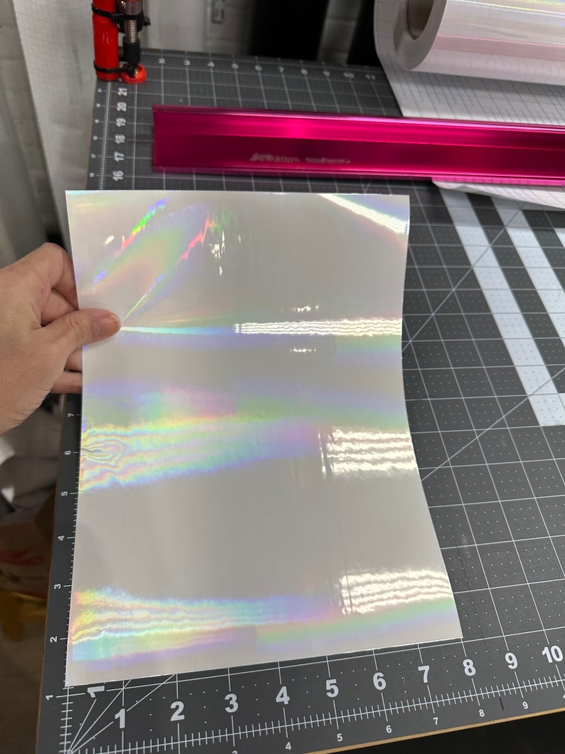 May include: A sheet of iridescent white vinyl with a holographic finish. The vinyl is lying flat on a black cutting mat with a ruler.
