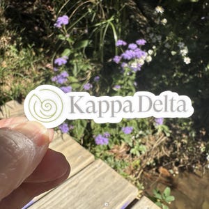 May include: A white sticker with the text "Kappa Delta" in gray lettering. The sticker has a green spiral design on the left side. The sticker is held up against a background of flowers and wood.