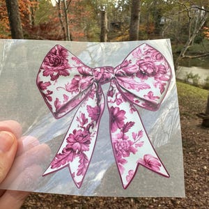May include: A large pink and white floral bow with a white background and pink floral pattern. The bow is on a clear plastic sheet.