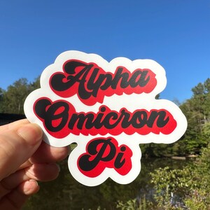 May include: A white sticker with red and black lettering that reads "Alpha Omicron Pi". The sticker is held in a hand against a blue sky background.