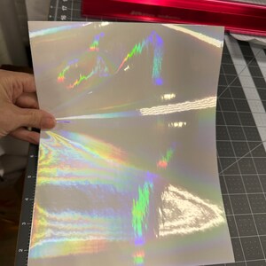 May include: A sheet of iridescent holographic vinyl with a rainbow pattern. The vinyl is white with a subtle shimmer and has a holographic effect that changes color depending on the angle of light.