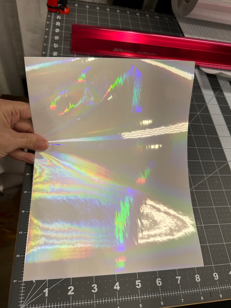 May include: A sheet of iridescent holographic vinyl with a rainbow pattern. The vinyl is white with a subtle shimmer and has a holographic effect that changes color depending on the angle of light.