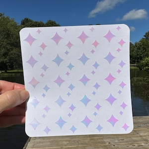 May include: A white square with rounded corners featuring a pattern of iridescent diamond shapes in shades of pink, purple, and blue. The design is held up against a backdrop of a lake and trees.