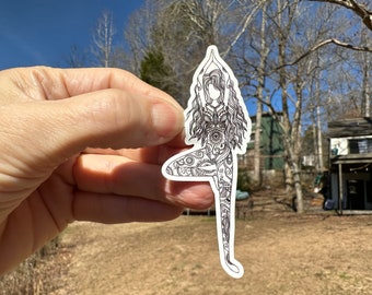 May include: A black and white sticker featuring a detailed illustration of a person in a yoga tree pose. The figure is adorned with intricate patterns and designs, with arms raised overhead. The sticker is held against a backdrop of a sunny outdoor scene.