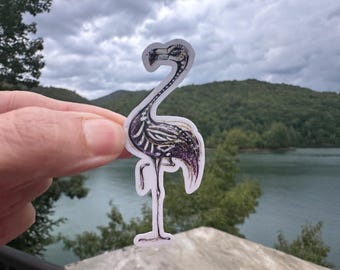 May include: A sticker featuring a flamingo skeleton design. The flamingo is depicted in black and white with purple accents, held against a backdrop of a lake and mountains under a cloudy sky.
