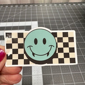 May include: A sticker with a green smiley face with fangs on a black and white checkered background.