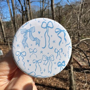 May include: A white, round button featuring a repeating pattern of light blue bows, hearts, and stars. The bows vary in style, with some having long ribbons. The background is a natural outdoor scene with trees.
