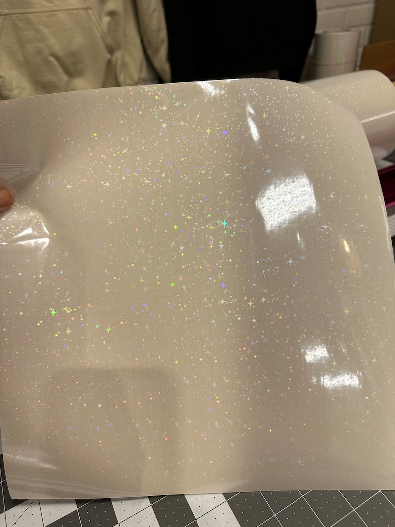 May include: A sheet of white glitter vinyl with a sparkling, iridescent finish. The vinyl is covered in tiny, star-shaped glitter particles that catch the light, creating a shimmering effect. The material is slightly curved, suggesting it is flexible.