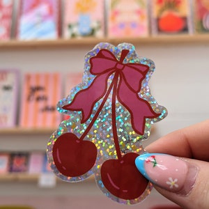 May include: A holographic sticker featuring a pair of red cherries with a pink bow. The sticker has a sparkly, iridescent finish.
