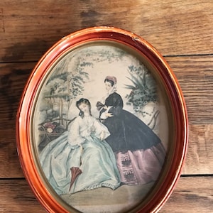 May include: An oval framed print of two women in a garden setting. The women are wearing long dresses and are holding a parasol and a book. The print is in shades of blue, green, pink, and black.