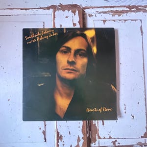 以下が含まれることがあります： サウスサイド・ジョニーのポートレートが描かれた、ビンテージのビニールレコードアルバムのカバー。カバーには「サウスサイド・ジョニー・アンド・ザ・アズベリー・ジュークス」と「ハーツ・オブ・ストーン」の文字が書かれている。