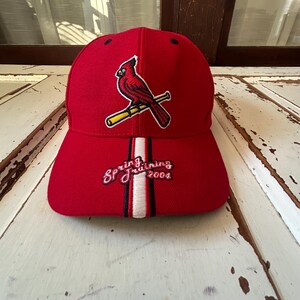 May include: Red baseball cap featuring a cardinal bird logo on the front. The cap has a curved brim and the text "Spring Training 2004" embroidered on the front. The cap has a white and black stripe design.