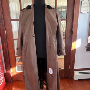 Puede incluir: Un abrigo tipo trench marr&oacute;n expuesto en un maniqu&iacute; negro. El abrigo tiene un dise&ntilde;o de doble botonadura con botones grandes, cuello con solapas y detalles de cintur&oacute;n en los pu&ntilde;os. El abrigo es largo, llegando a los tobillos.