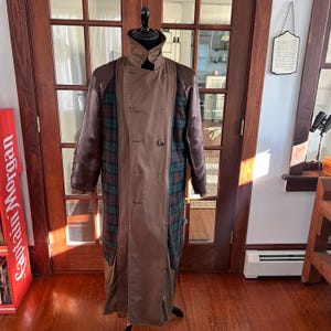 Puede incluir: Un abrigo tipo trench marr&oacute;n con cuello alto y dise&ntilde;o cruzado. El abrigo presenta paneles a cuadros en tonos verdes, azules y rojos. Las mangas son de un marr&oacute;n m&aacute;s oscuro y el abrigo se exhibe en un maniqu&iacute;.