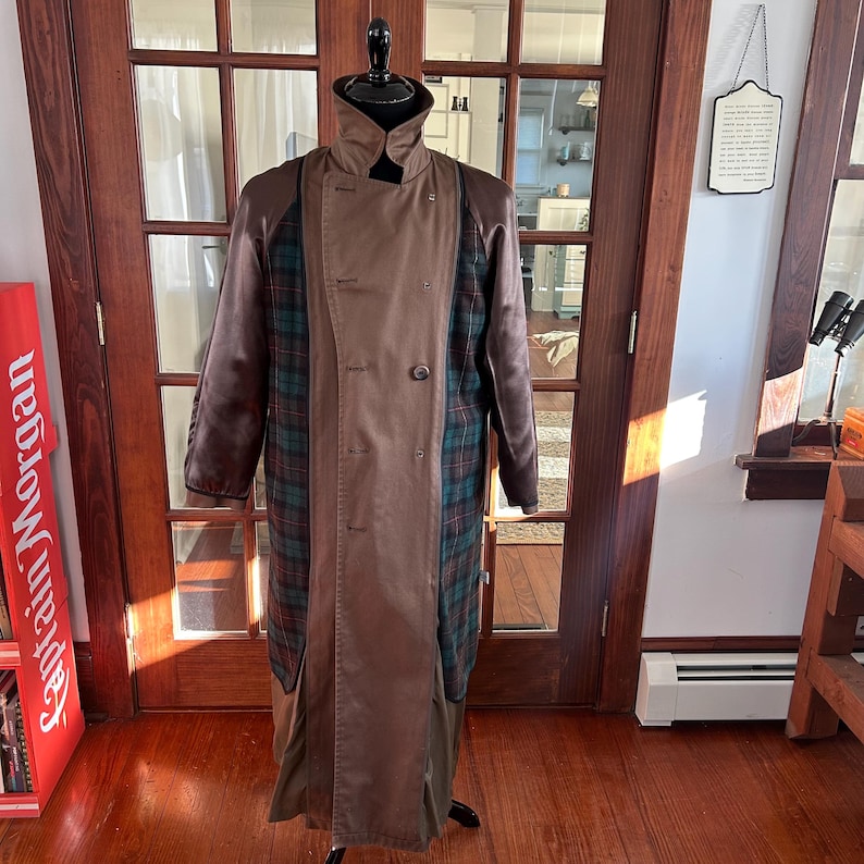 Puede incluir: Un abrigo tipo trench marr&oacute;n con cuello alto y dise&ntilde;o cruzado. El abrigo presenta paneles a cuadros en tonos verdes, azules y rojos. Las mangas son de un marr&oacute;n m&aacute;s oscuro y el abrigo se exhibe en un maniqu&iacute;.