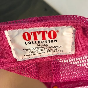 Op de afbeelding: Roze mesh trucker cap met een wit label met de tekst "OTTO COLLECTION 32-468 100% Polyester 100% Nylon One Size Fits Most Made in China RN 129002"