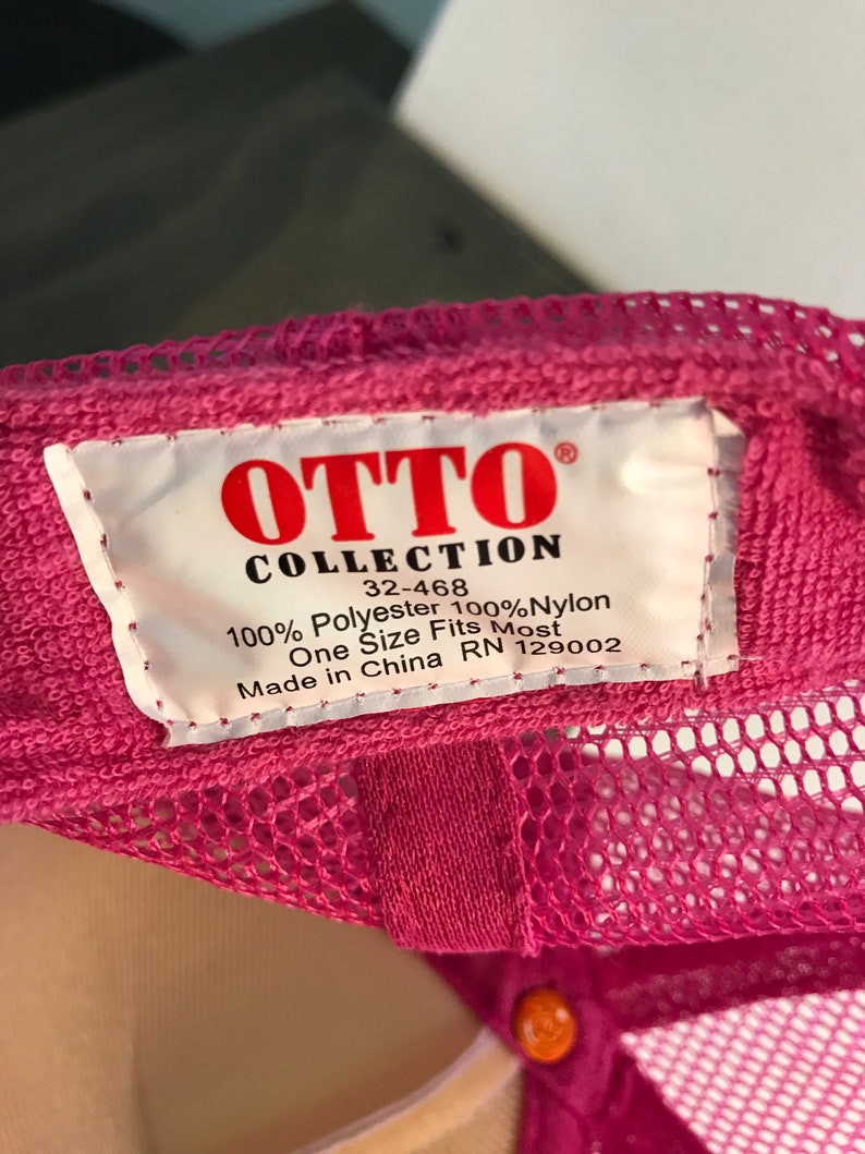 Op de afbeelding: Roze mesh trucker cap met een wit label met de tekst "OTTO COLLECTION 32-468 100% Polyester 100% Nylon One Size Fits Most Made in China RN 129002"