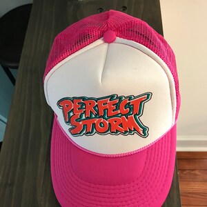 Op de afbeelding: Een roze en witte truckercap met een mesh achterkant. De voorkant van de cap heeft een witte patch met de tekst "PERFECT STORM" in oranje, groene en witte letters.