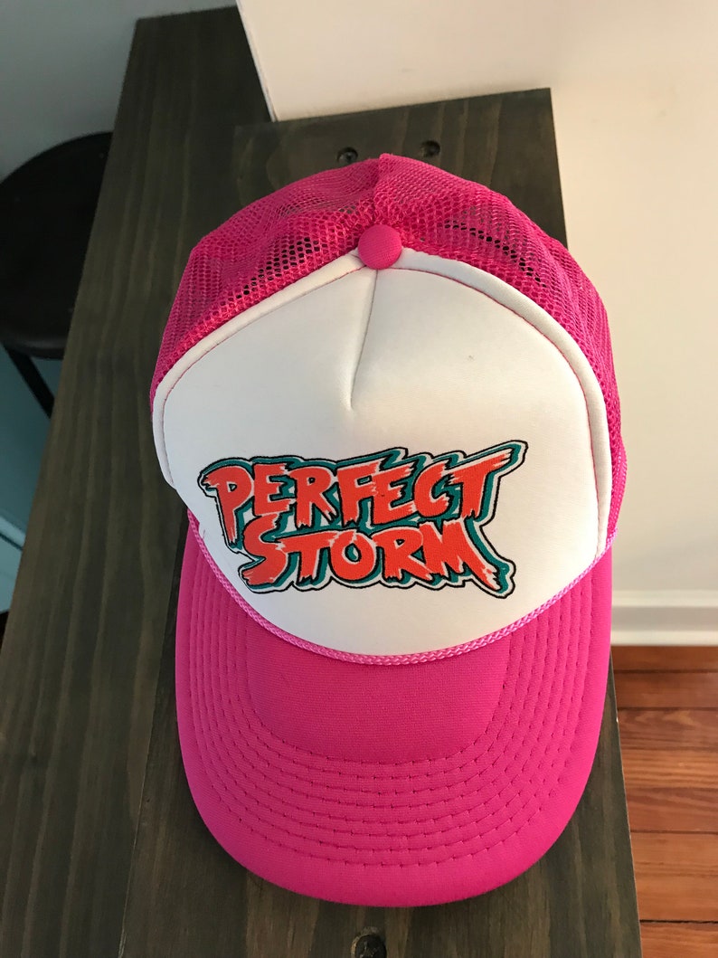 Op de afbeelding: Een roze en witte truckercap met een mesh achterkant. De voorkant van de cap heeft een witte patch met de tekst "PERFECT STORM" in oranje, groene en witte letters.