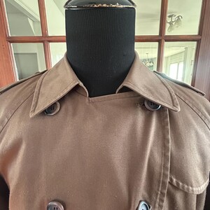 Puede incluir: Un abrigo tipo trench marr&oacute;n con un dise&ntilde;o cruzado. El abrigo presenta un cuello con muescas, charreteras y botones grandes y oscuros. El abrigo se exhibe en una cabeza de maniqu&iacute; negra.