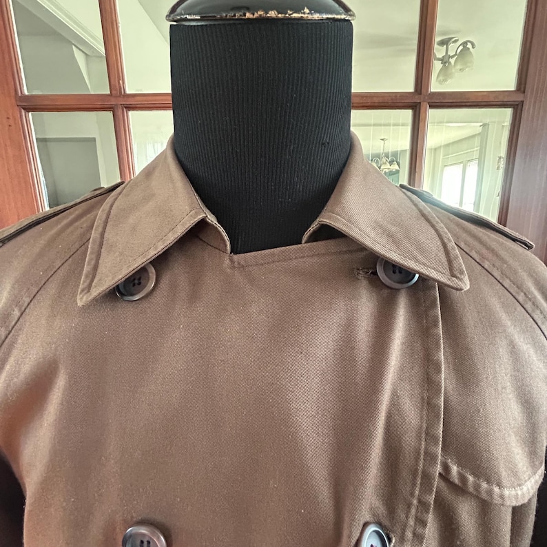 Puede incluir: Un abrigo tipo trench marr&oacute;n con un dise&ntilde;o cruzado. El abrigo presenta un cuello con muescas, charreteras y botones grandes y oscuros. El abrigo se exhibe en una cabeza de maniqu&iacute; negra.