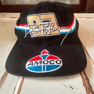 Puede incluir: Gorra de béisbol negra con un número 95 dorado y el texto "Rare Blanc" en blanco. La gorra también tiene un diseño de rayas rojo, blanco y azul y un logotipo Amoco azul, blanco y rojo.