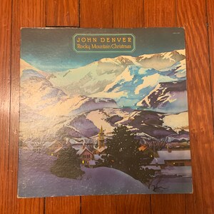 Puede incluir: Una portada de disco de vinilo cuadrada con una ilustración estilo acuarela de un pueblo de montaña cubierto de nieve al atardecer. El texto John Denver y Rocky Mountain Christmas está impreso en un marco decorativo sobre un cielo azul.