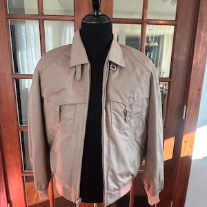 Op de afbeelding: Een bruinbeige bomberjack met ritssluiting, twee voorzakken en een kraag. De jas wordt getoond op een zwarte mannequin. De manchetten en zoom zijn een lichtere tint bruinbeige.