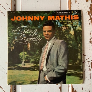 May include: A vintage vinyl record album cover featuring Johnny Mathis. The album cover is green and features a photo of Johnny Mathis in a grey suit and red tie. The album title is "Swing Softly" and the text "Orchestra Under The Direction Of Percy Faith" is also included.
