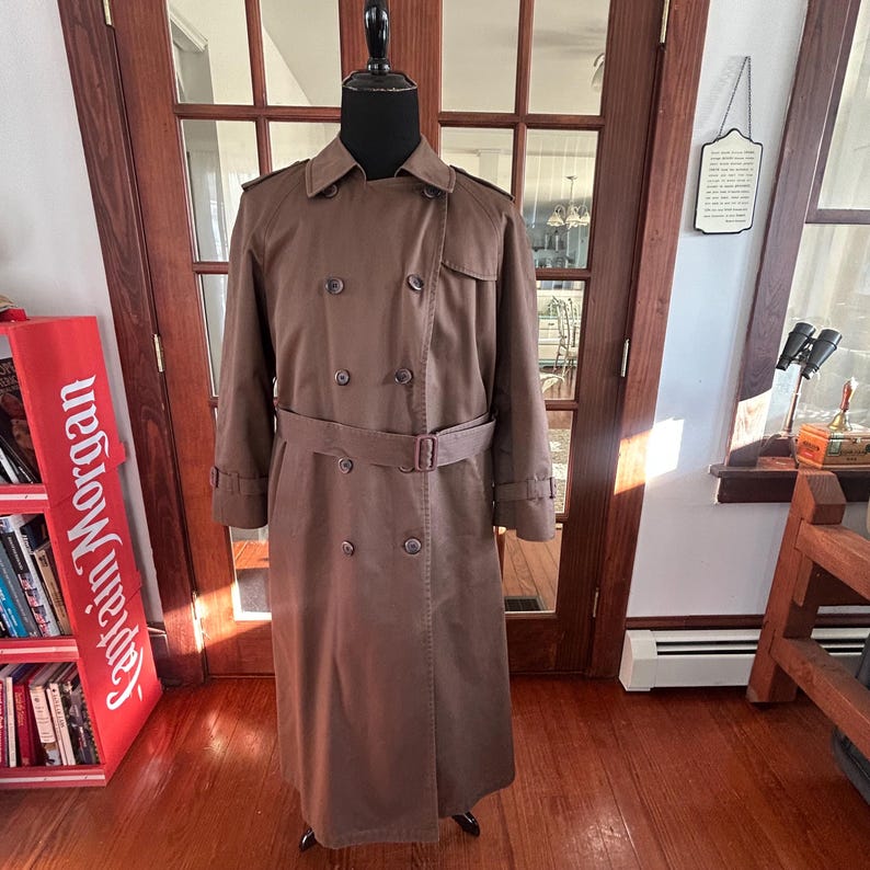 Puede incluir: Un abrigo tipo gabardina marr&oacute;n con dise&ntilde;o cruzado, cintur&oacute;n y botones. El abrigo se exhibe en un maniqu&iacute;. Es un estilo cl&aacute;sico, adecuado para diversas ocasiones.