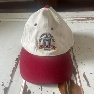 Op de afbeelding: Een crèmekleurige baseballpet met een bordeauxrode klep en knoop. De pet heeft een geborduurd embleem met de tekst "OLYMPIA FIELDS U.S. OPEN 2003". Deze pet is waarschijnlijk een souvenir of verzamelobject.