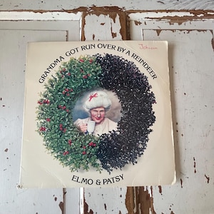 May include: A vintage vinyl record album cover featuring a woman in a red and white Santa hat holding a cup of coffee. The album cover is titled "Grandma Got Run Over By A Reindeer" by Elmo & Patsy. The album cover is signed "Johnson".