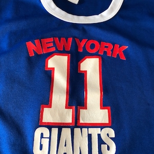 May include: Blue and white t-shirt with the text "NEW YORK 11 GIANTS" in red and white lettering.