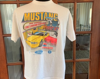 Op de afbeelding: Wit T-shirt met een afbeelding van een gele Ford Mustang en een rode Ford Mustang. De afbeelding bevat de tekst "Mustang", "Now Showing", "The Legend Continues", "30th Anniversary Celebration", "Charlotte Motor Speedway" en "Ford".