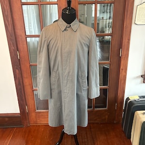 May include: A grey long-sleeved coat with a button fastening and two pockets. The coat is on a mannequin in front of a wooden door.